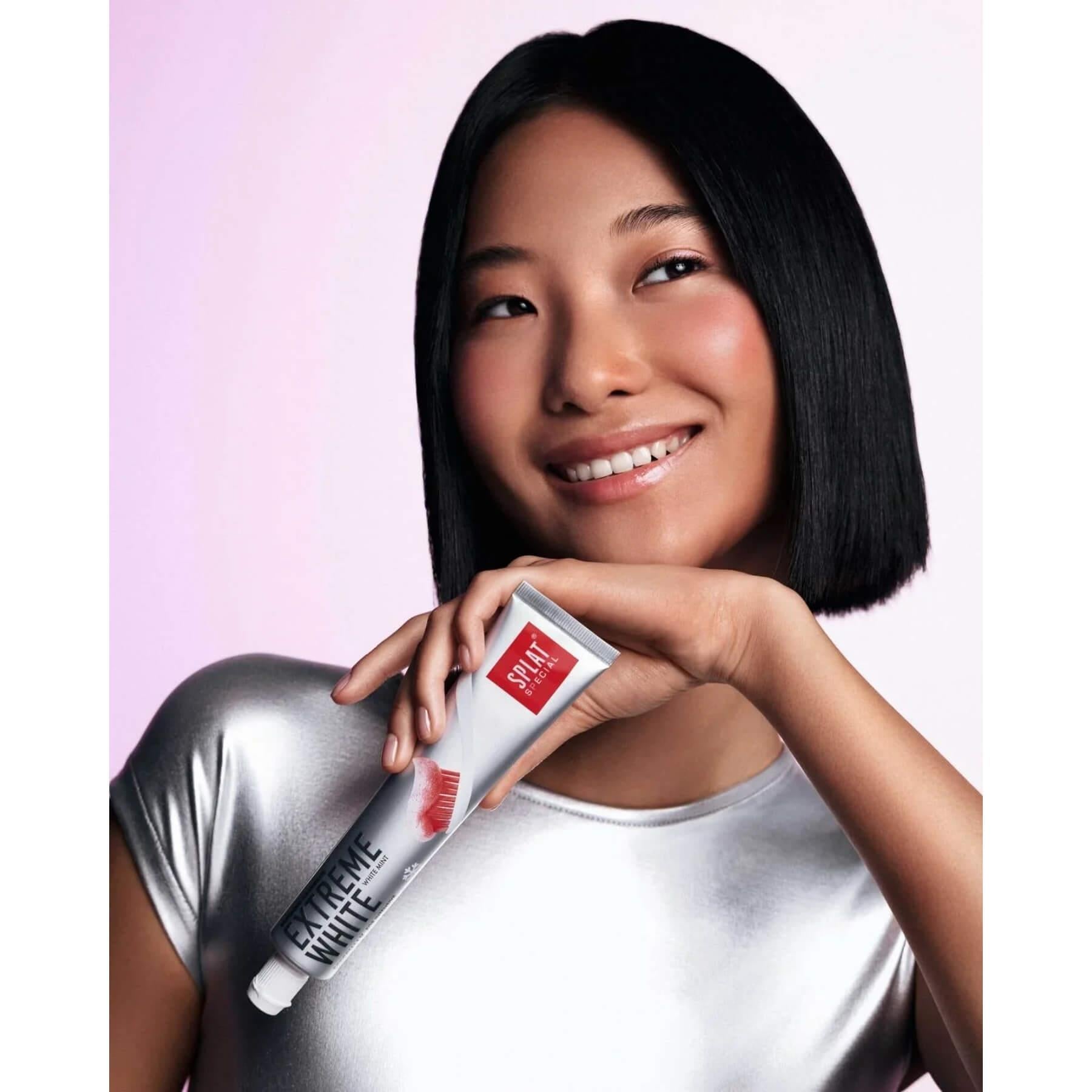 A smiling woman with a short black bob holds a tube of Style hair color against a soft pink studio background.