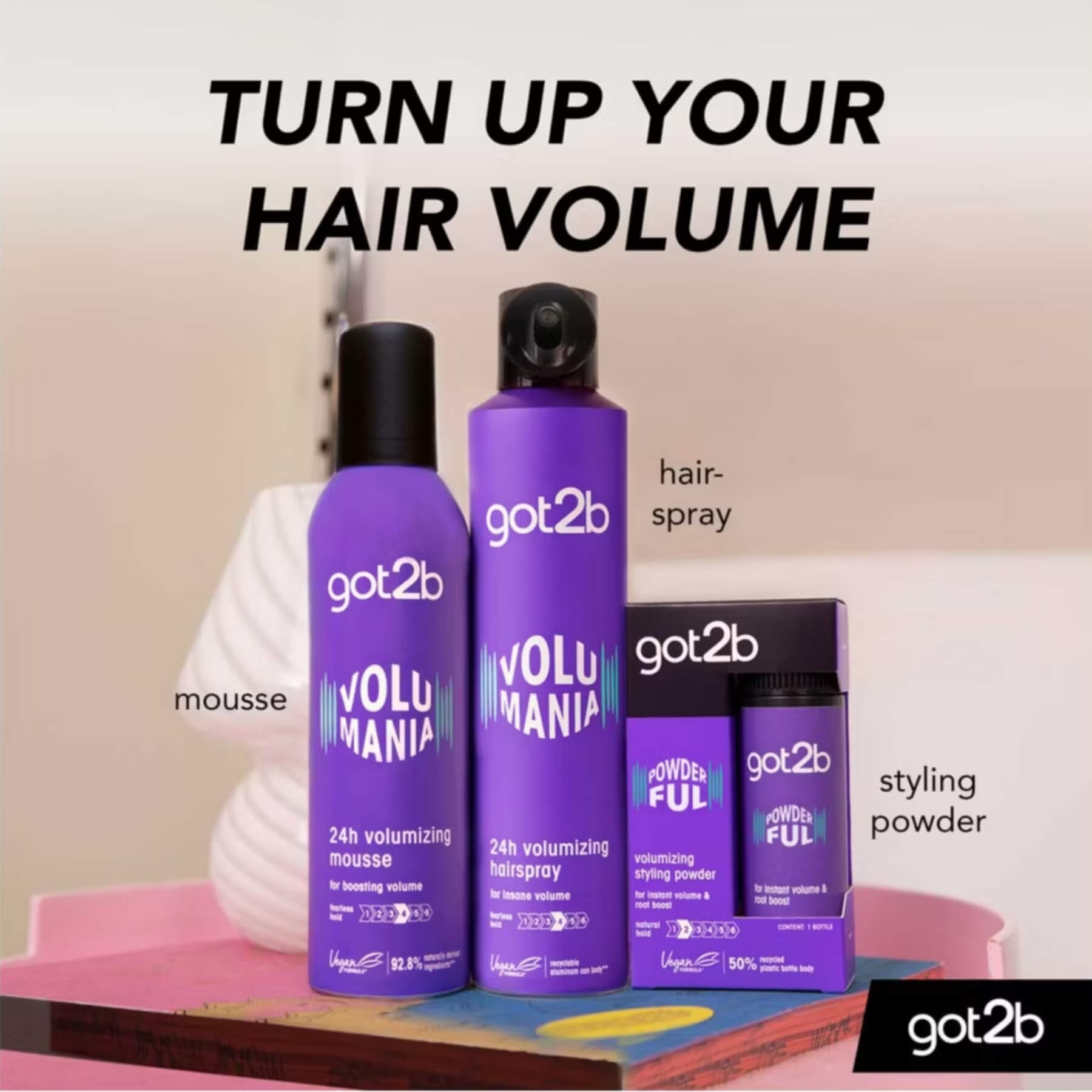 A promotional product shot for got2b Volumania hair products on a pastel studio background. Visible items include a purple mousse bottle, a taller purple hair spray can, a small purple styling powder jar, and a black powder box, with text labels pointing to each product and the headline 'TURN UP YOUR HAIR VOLUME'.
