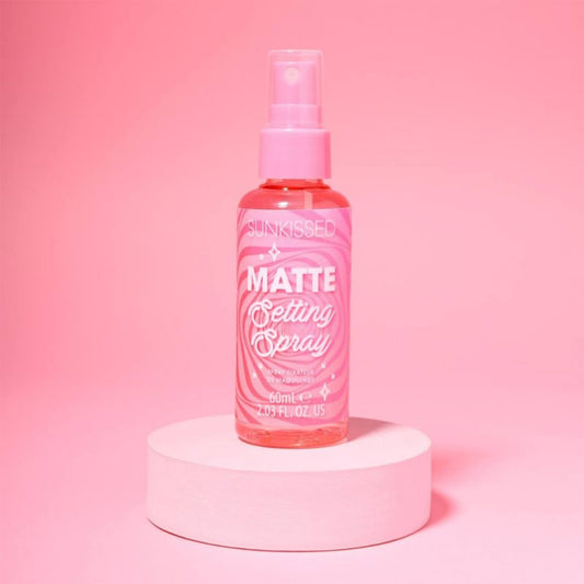 A pink bottle of Sunkissed Matte Setting Spray with a matching pink pump, displayed on a round pink pedestal against a solid pink background.