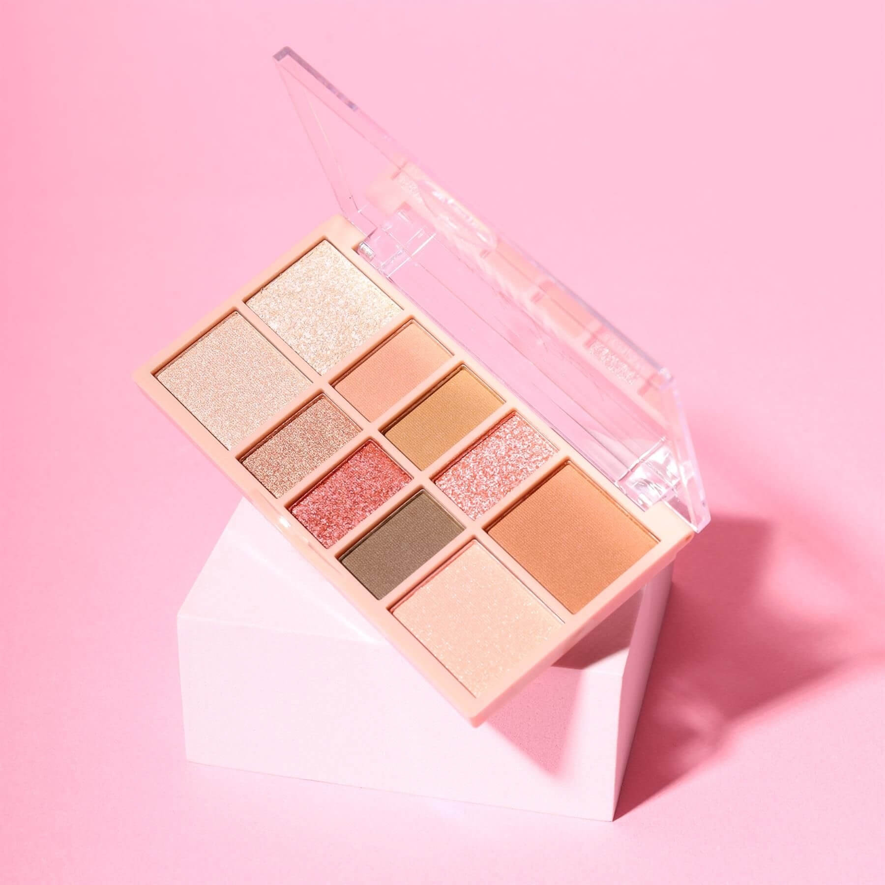An open 10-shade eyeshadow palette in a peach-toned case, displayed on a white geometric pedestal against a solid pink background.