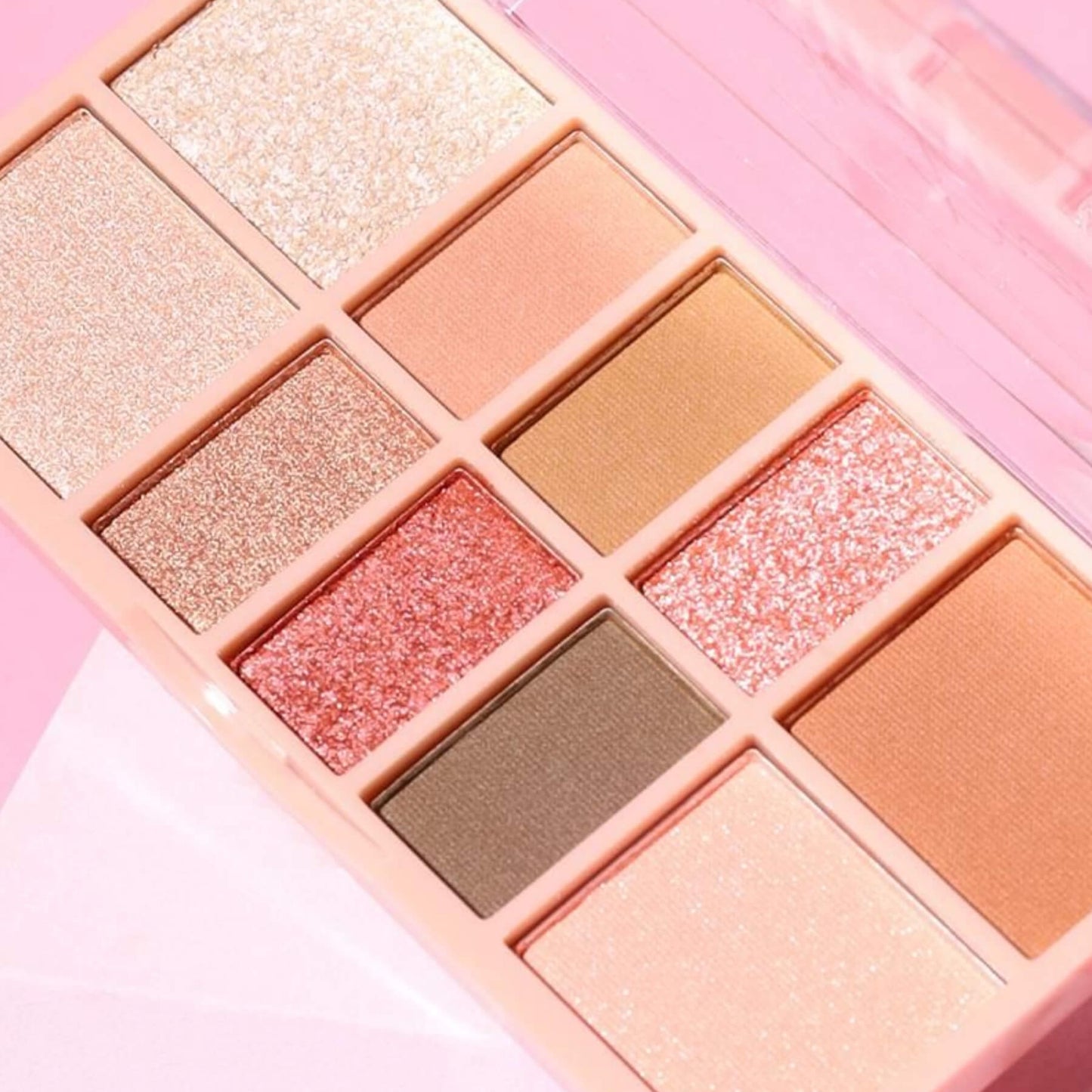 A close-up of a 12-pan eyeshadow palette in a pastel pink case, featuring various shimmery and matte shades in gold, bronze, rose gold, copper, and brown tones.