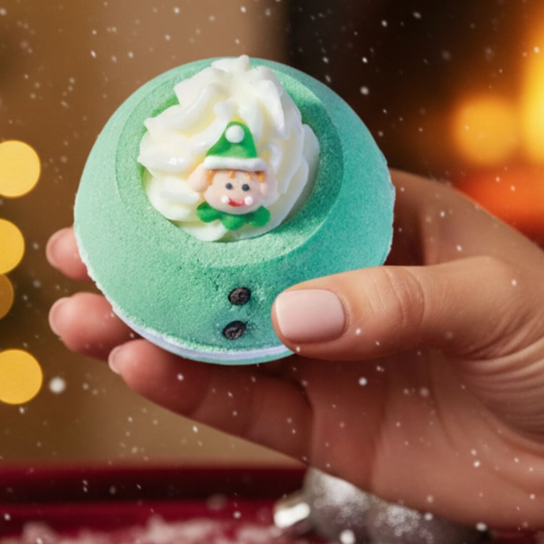Hand holding a green bath bomb with a snowman decoration against a warm, blurred background.